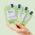 Woman’s hand holding 4 sachets from Pedi Moments Green Tea Detox on a green background.