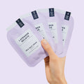 Woman’s hand holding 4 sachets from Pedi Moments Lavender Relieve on a purple background.