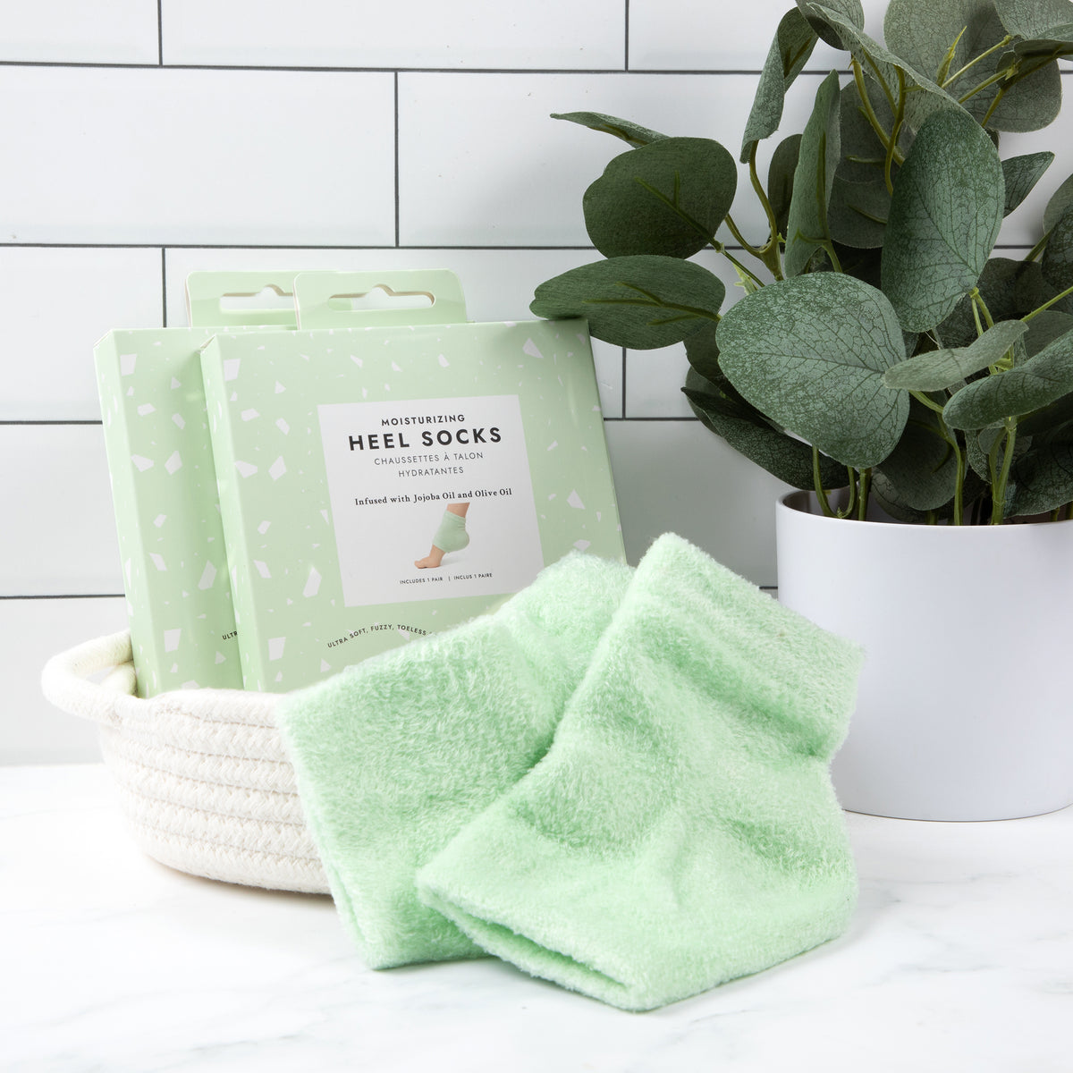2 Moisturizing heel sock boxes in a basket on a white countertop with subway tiles behind it sitting next to a green plant and the socks sitting in front of them