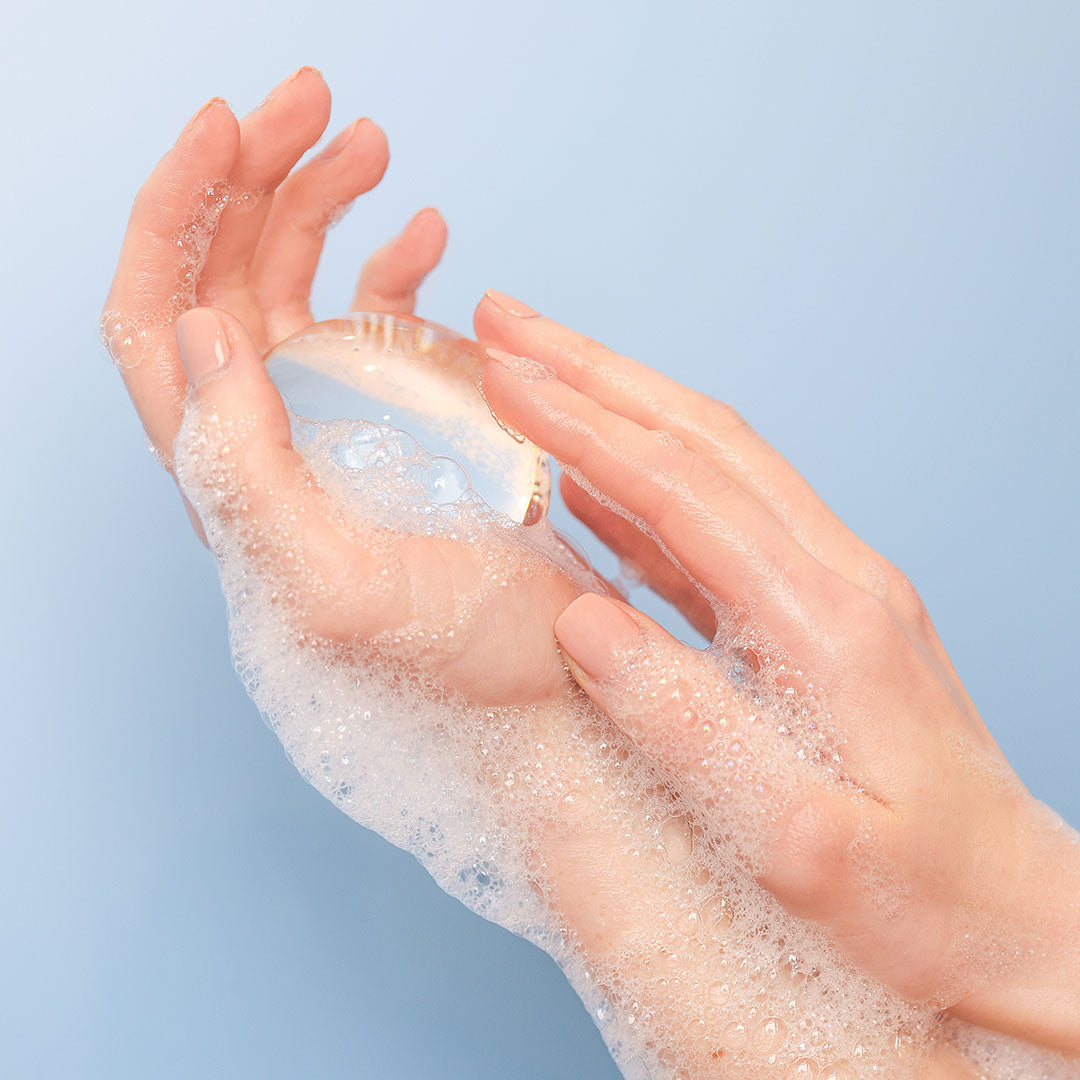 Woman’s hands holding Crystal Clear Head-To-Toe Cleansing Soap in a soapy lather on a blue background.