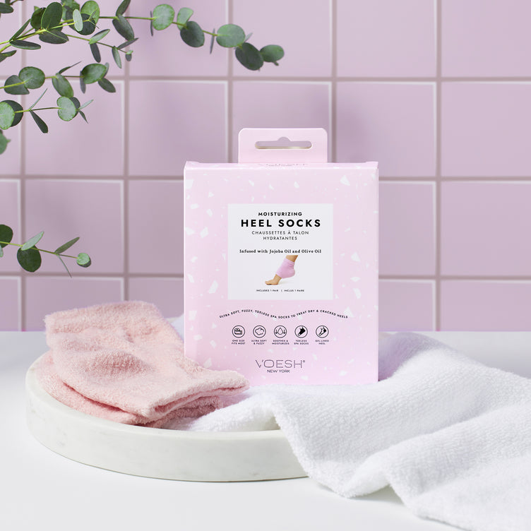 Pink Moisturizing Heel Socks next to packaging and a white hand towel on top of a white vanity tray on a pink tile background.