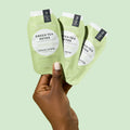 Woman’s hand holding 3 sachets from Mani Moments Green Tea Detox on a green background.
