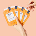 Woman’s hand holding 4 sachets from Pedi Moments Tangerine Glow on an orange background.