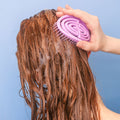 Woman using Scalp Massager in soapy hair pictured on a blue background.