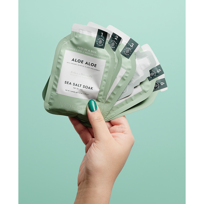 Woman's hand holding aloe aloe pedi in a box 6 step packets against a green background