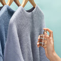 Woman spraying Room & Fabric Fragrance on sweaters pictured on a blue background.