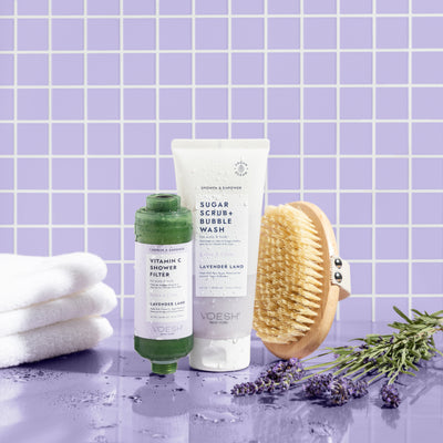 Lavender Land Shower & Empower Duo with a dry body brush, white towel, and lavender flowers on a purple tiled background.