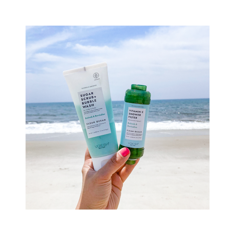 Woman's hand holding clean ocean sugar scrub and shower filter against a beach ocean scene