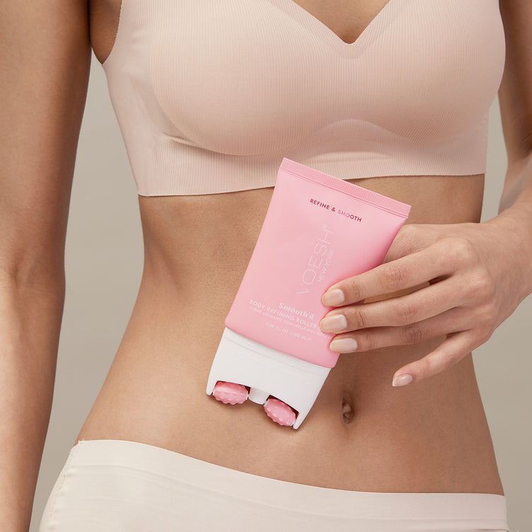 Woman in her bra and underwear holding Smooth’d Body Refining Roller Crème in front of her stomach pictured on a tan background.Woman in her bra and underwear holding Smooth’d Body Refining Roller Crème in front of her stomach pictured on a tan background.
