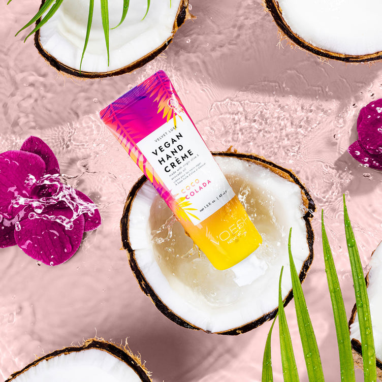 Coco Colada hand and body creme in a coconut floating in water against pink background with tropical flowers, palm leaves, and coconut halves