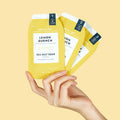Woman's hand holding lemon quench pedi in a box 3 step packets on yellow background